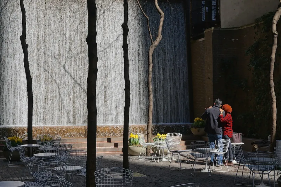 Man and woman watch waterfall in Paley Pocket Park in Manhattan, New York City. Park is filled with white tables and chairs.
